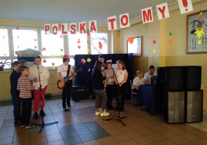 Uczniowie stoją przy mikrofonach i śpiewają w szkolnym chórze. Na środku stoi uczeń z gitarą i gra. Nad uczniami wisi czerwono-biały napis Polska to my!